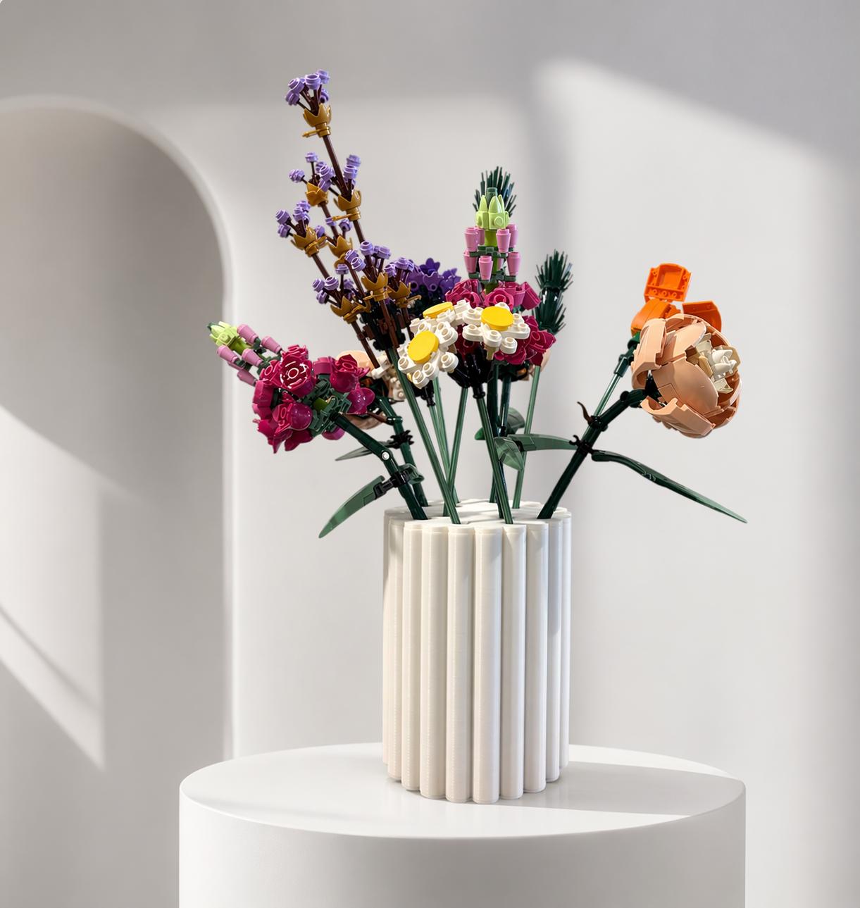 May include: A white, fluted vase holds a colorful arrangement of artificial flowers. The bouquet includes purple lavender, pink and white daisies, and an orange peony. The vase sits on a white pedestal against a soft, neutral background.