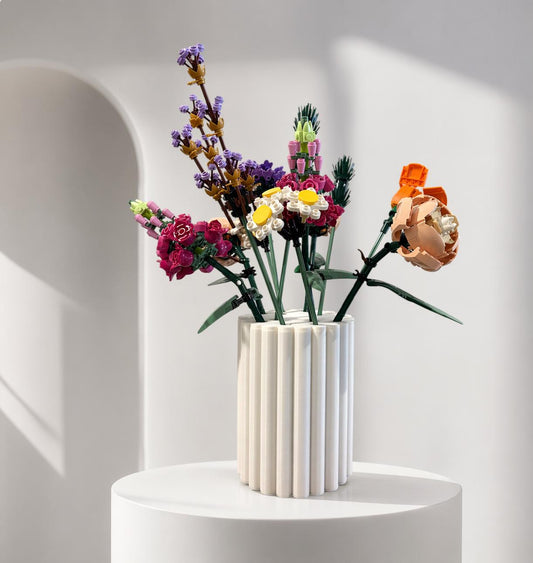 May include: A white, fluted vase holds a colorful arrangement of artificial flowers. The bouquet includes purple lavender, pink and white daisies, and an orange peony. The vase sits on a white pedestal against a soft, neutral background.