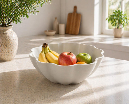 May include: A white, scalloped-edge ceramic fruit bowl filled with fresh fruit. The bowl contains bananas, a red apple, a green apple, and an orange. The bowl sits on a light-colored countertop, with a vase of greenery in the background.