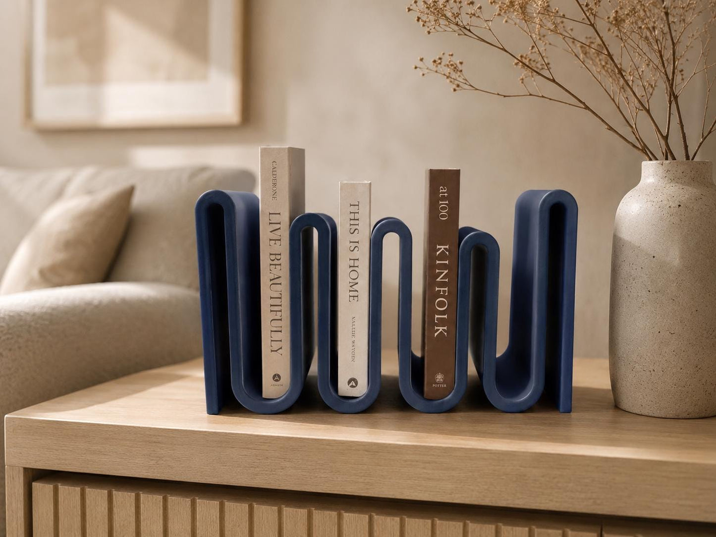 May include: A navy blue, wavy-shaped bookend holds three books with titles "LIVE BEAUTIFULLY", "THIS IS HOME", and "KINFOLK". A beige vase with dried flowers sits to the right. The bookend is on a light wood surface.