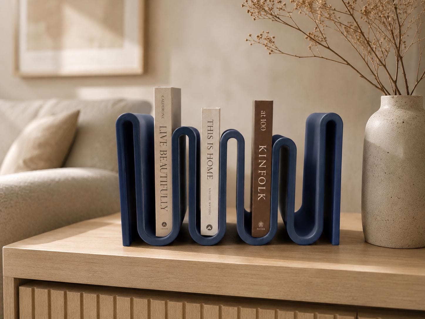 May include: A navy blue, wavy-shaped bookend holds three books with titles "LIVE BEAUTIFULLY", "THIS IS HOME", and "KINFOLK". A beige vase with dried flowers sits to the right. The bookend is on a light wood surface.