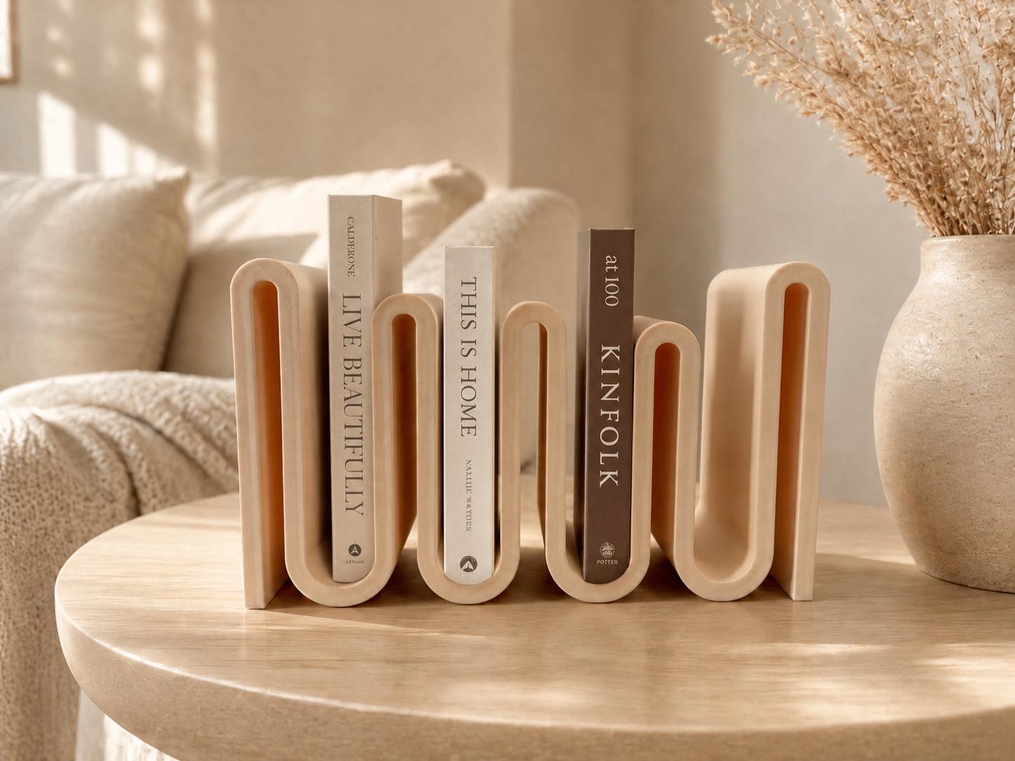 May include: A beige, wavy-shaped bookend holds three books with titles including "LIVE BEAUTIFULLY", "THIS IS HOME", and "KINFOLK". The bookend sits on a light-colored wooden table, with a vase of dried flowers in the background.